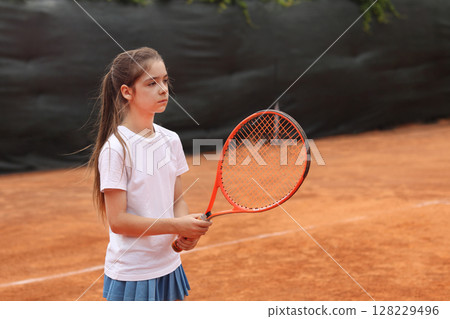 Young girl holding a tennis racket on a clay court, ready to play Young girl holding a tennis racket on a clay court, ready to play 128229496