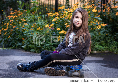 Young girl sitting with a skateboard near a garden of yellow flowers Young girl sitting with a skateboard near a garden of yellow flowers 128229560
