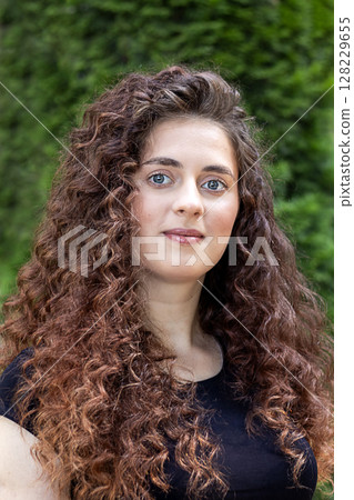Young woman with curly hair posing against a lush green backdrop Young woman with curly hair posing against a lush green backdrop 128229655