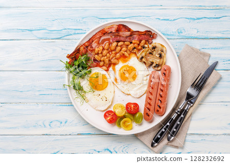 A classic breakfast plate featuring sunny-side-up eggs, crispy bacon, sausages, and fresh tomatoes 128232692
