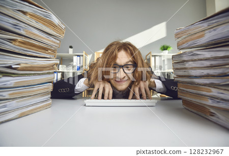 Happy office worker sitting at desk with stacks of papers and typing on wireless keyboard 128232967
