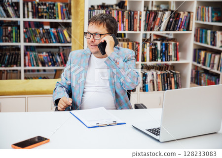 Business man talking on phone working on laptop online in library Business man talking on phone working on laptop online in library 128233083