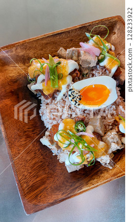 A wooden plate filled with brown rice, topped with tofu cubes, halved boiled egg and various colorful garnishes, creating a fresh and vibrant look for a healthy vegetarian meal 128233922