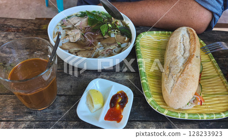 A casual authentic traditional local Vietnamese meal with a bowl of pho bo or beef noodle soup, iced coffee, and a baguette sandwich or banh mi on a rustic wooden table 128233923