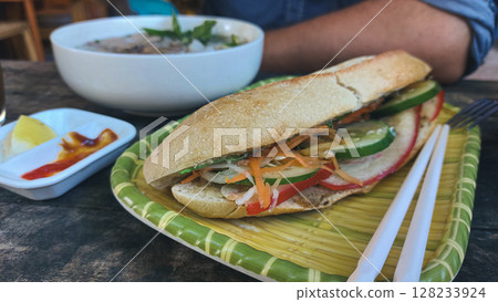 Close up to a baguette sandwich or banh mi on a plate set on a wooden table with a bowl of pho bo and a person in the background showing the authentic traditional local Vietnamese cuisine Close up to a baguette sandwich or banh mi on a plate set on a wooden table with a bowl of pho bo and a person in the background showing the authentic traditional local Vietnamese cuisine 128233924
