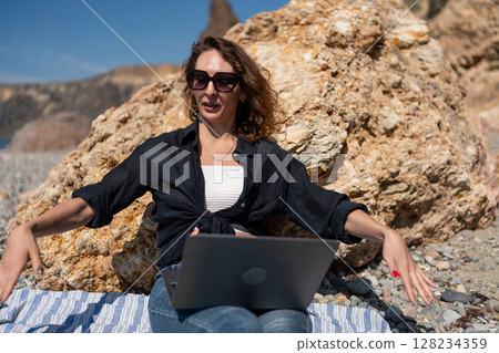 Laptop beach woman: Sunny day; Woman works remotely on laptop beside rock formation at beach. Laptop beach woman: Sunny day; Woman works remotely on laptop beside rock formation at beach. 128234359