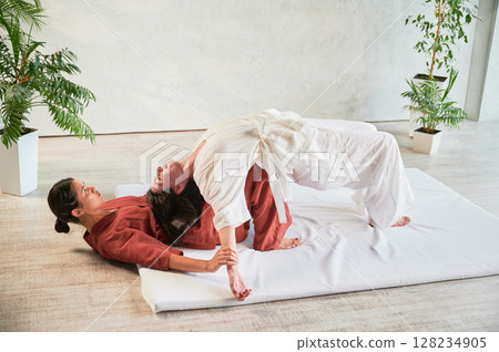 Masseuse making thai yoga massage. Woman therapist doing traditional massage treatment, stretching female patient's body. Client lying on mat, enjoying hand massage in calm atmosphere. 128234905