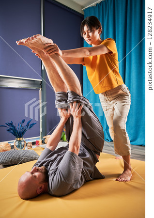 Masseuse making thai yoga massage. Woman therapist doing traditional massage treatment, stretching male patient's body. Bald client lying on mat, enjoying hand massage in calm atmosphere. 128234917