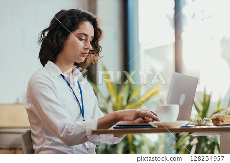 Young woman typing on laptop during lunch break in bright office cafeteria with coffee 128234957