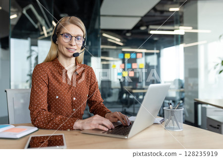 A smiling blonde woman wearing a headset works on a laptop in a modern office setting. She looks friendly and helpful. 128235919