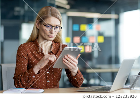 A businesswoman is working at her desk with a tablet and laptop in a modern office setting, wearing glasses and a button-down shirt. 128235926