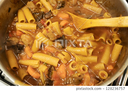 Chef preparing beef stew with rigatoni pasta and vegetables in a stainless steel pot 128236757