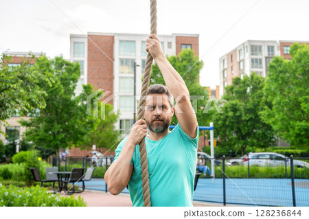 Fitness rope climb exercise in fitness gym workout outdoor. Strong man climbing on the rope during training in sport gym. Mature man athlete climbing a rope. Man do functional training Fitness rope climb exercise in fitness gym workout outdoor. Strong man climbing on the rope during training in sport gym. Mature man athlete climbing a rope. Man do functional training 128236844