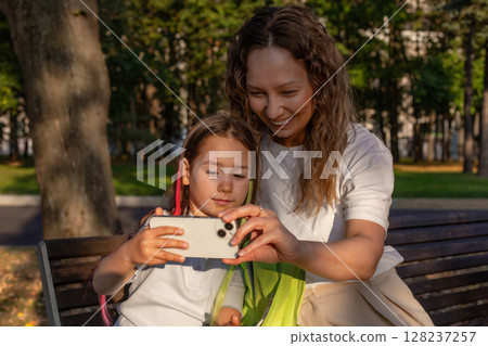 Mother and daughter enjoying a summer day taking selfies on a park bench 128237257