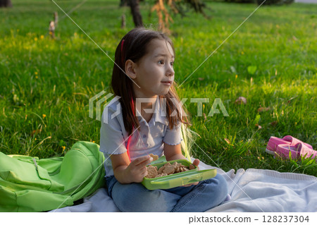 Little girl enjoying picnic lunch outdoors amidst greenery 128237304