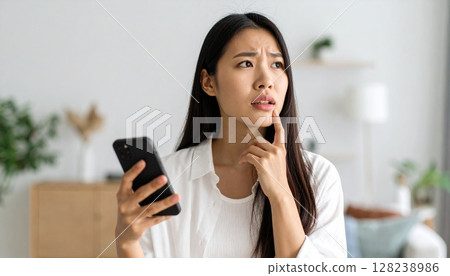An image of a woman holding a smartphone in her hand and looking worried or in distress. 128238986