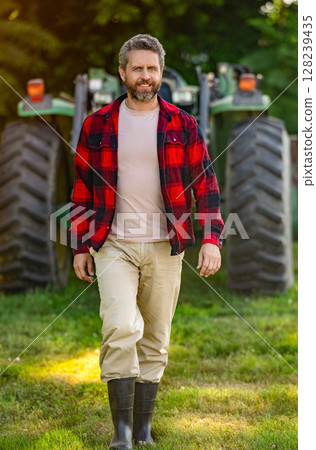 Farmer with harvesting machinery. Mature man at tractor. Tractor driver. Agriculture and field work. Farmer in field wearing gum boots with tractor outdoor. Crop harvesting. Agriculture machine 128239435