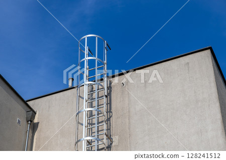 Tall silver industrial caged access ladder firmly attached to a plain grey concrete building wall, representing safety, emergency exit, and rooftop access solutions 128241512