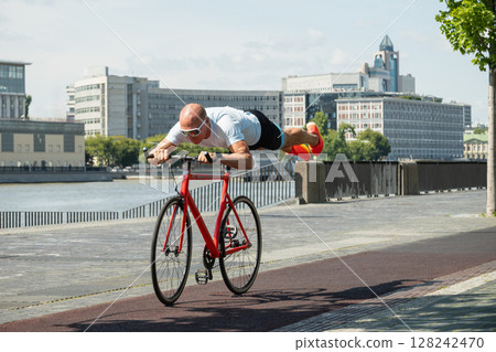 Happy cyclist rides bicycle at urban public park. Man riding bicycle, lying on seat with feet in air. Summer outdoor activity. Happy cyclist rides bicycle at urban public park. Man riding bicycle, lying on seat with feet in air. Summer outdoor activity. 128242470