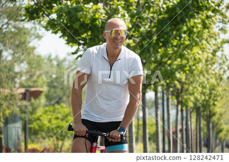 Happy cyclist smiling rides bicycle at urban public park. Sporty man wears reflective sunglasses. Summer outdoor activity. 128242471