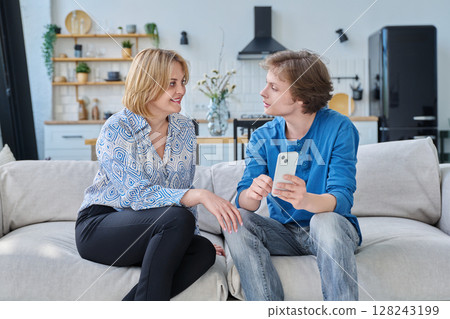 Middle aged mother and teenage son sitting together at home looking at smartphone Middle aged mother and teenage son sitting together at home looking at smartphone 128243199