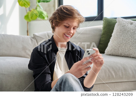 Happy joyful handsome teenager guy using smartphone sitting on floor at home. 128243209