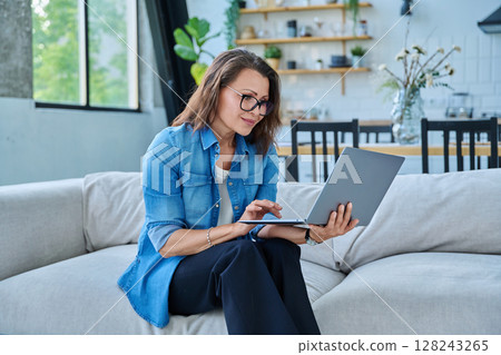 Portrait of middle aged woman using laptop sitting on sofa at home Portrait of middle aged woman using laptop sitting on sofa at home 128243265