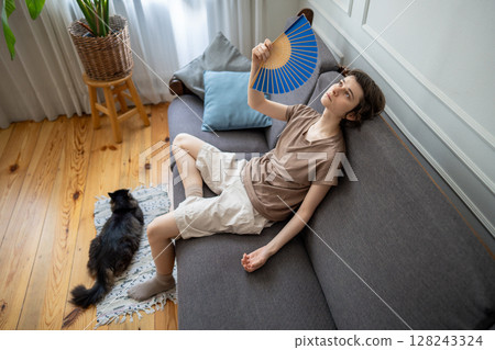 Pet owner and cat in summer heat. Exhausted woman fanning on sofa, pet lying in heat distress Pet owner and cat in summer heat. Exhausted woman fanning on sofa, pet lying in heat distress 128243324