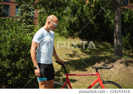 Serious cyclist ready to ride bicycle at urban public park. Sporty man wears reflective sunglasses. Summer outdoor activity. Serious cyclist ready to ride bicycle at urban public park. Sporty man wears reflective sunglasses. Summer outdoor activity. 128243418