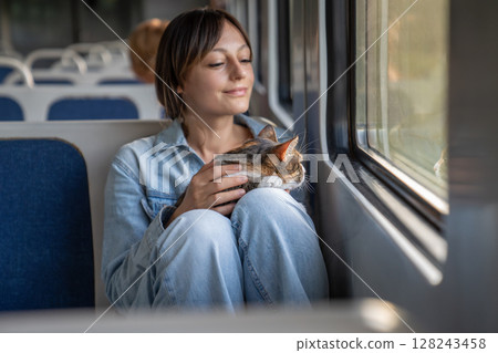 Public train ride of thoughtful smiling woman with calm peaceful tricolor cat sitting on lap. 128243458