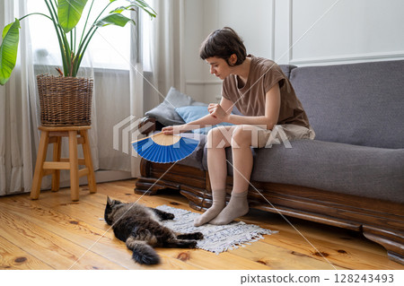 Caring woman pet owner gently fanning cat curling on floor to ease heat stress at home, no AC 128243493