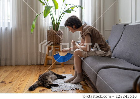 Caring woman pet owner gently fanning cat curling on floor to ease heat stress at home, no AC 128243516