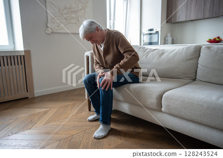 Tired exhausted senior man grimacing while attempting to stand with knee joint destruction from sofa Tired exhausted senior man grimacing while attempting to stand with knee joint destruction from sofa 128243524