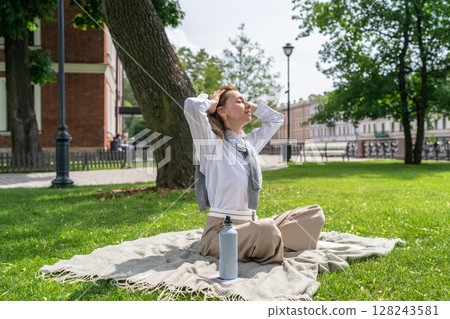 Calm woman open face to sun while sitting on lawn in sunny summer park, sun protection, UV heat Calm woman open face to sun while sitting on lawn in sunny summer park, sun protection, UV heat 128243581