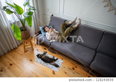 Lazy bored woman lounging on sofa scrolling through smartphone web content as cat rests close by 128243659