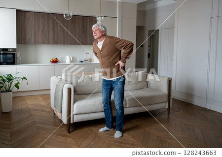 Hunched suffering elderly pensioner clutches painful lower back near sofa at home alone, hernia Hunched suffering elderly pensioner clutches painful lower back near sofa at home alone, hernia 128243663