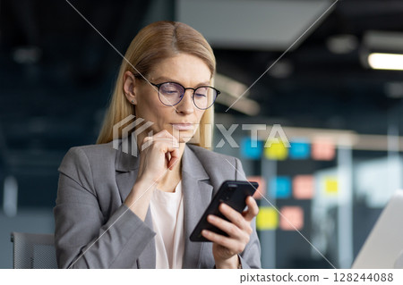 A thoughtful businesswoman in a gray suit is focused on her smartphone, with a hand on her chin. A thoughtful businesswoman in a gray suit is focused on her smartphone, with a hand on her chin. 128244088
