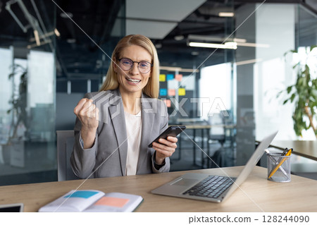 A businesswoman celebrates success at her desk, holding a phone and laptop, in an office environment. 128244090