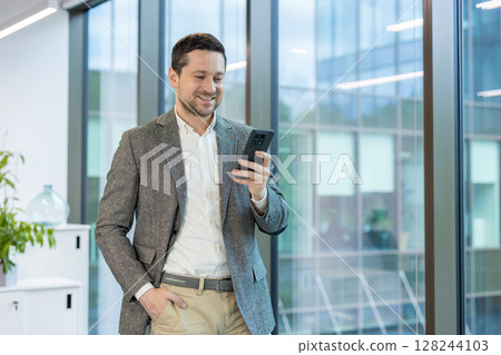 Smiling young male businessman standing in office, holding hand in pocket and using mobile phone. 128244103