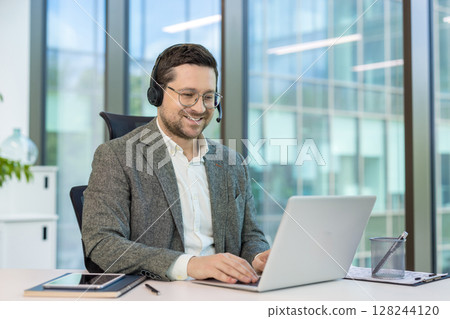 Smiling young office worker in headset sitting at desk in office and working on laptop. Smiling young office worker in headset sitting at desk in office and working on laptop. 128244120