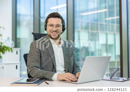 Portrait of a smiling young man in a suit and headset sitting at a working office desk and looking at the camera. 128244121