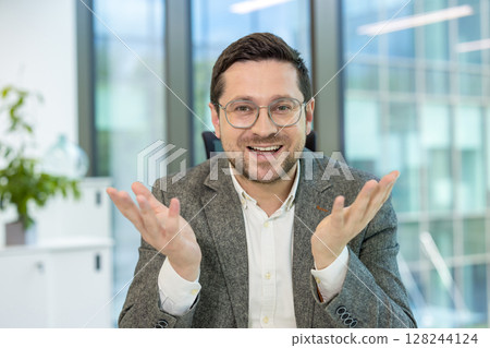 A young businessman in a suit and glasses sits in the office at a desk and talks to the camera while gesturing with his hands. 128244124