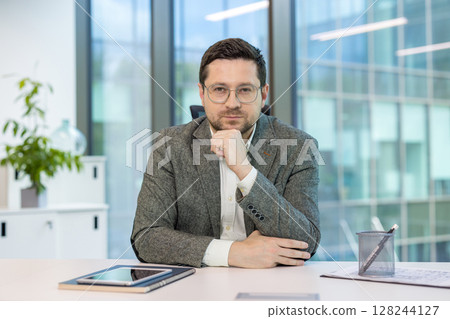 A serious and successful company CEO and director sits at a desk in an office in a suit, holds his hand near his face, and looks confidently into the camera. 128244127
