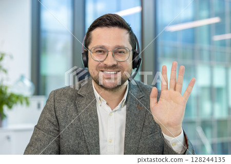 Close-up portrait of a young man in a suit and headset sitting at a desk in an office, looking and waving at the camera. 128244135