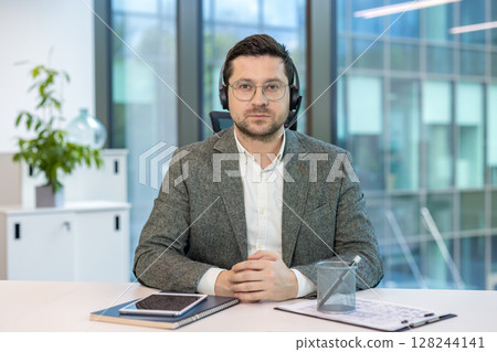 Close-up portrait of a smiling young man in a headset and suit sitting at a desk in the office, talking and waving at the camera. Close-up portrait of a smiling young man in a headset and suit sitting at a desk in the office, talking and waving at the camera. 128244141