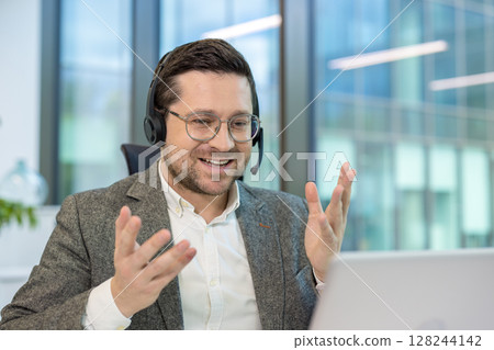 A young male office worker sits at a desk in the office, wearing a headset and talking on a video call on a laptop, gesturing with his hands. A young male office worker sits at a desk in the office, wearing a headset and talking on a video call on a laptop, gesturing with his hands. 128244142