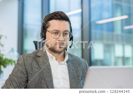 Close-up photo of a young man in a suit and headset working seriously in the office on a laptop. 128244145