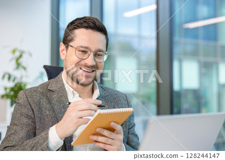 Close-up photo of a young male office worker sitting in front of a laptop screen, chatting online and taking notes in a notebook. Close-up photo of a young male office worker sitting in front of a laptop screen, chatting online and taking notes in a notebook. 128244147