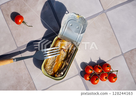 Tin of sardine with fork and tomato on gray tile. Open canned fish for quick dish concept. 128244163