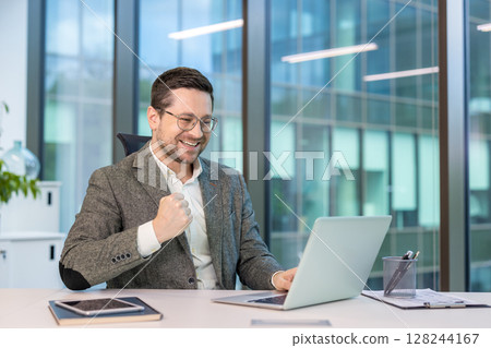 Happy young businessman sitting at a desk in the office, looking at the laptop screen and rejoicing at the news and smiling, showing a victory gesture with his hand. 128244167